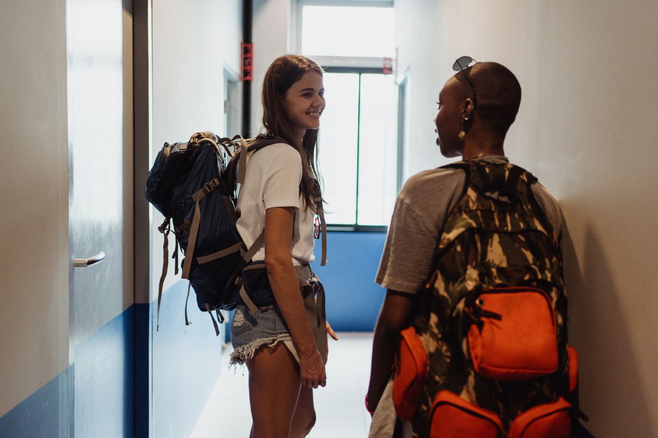 About Two women travelers chatting in a hostel hallway, smiling and carrying backpacks.