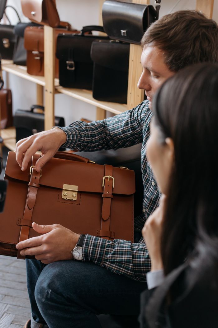 A man sits indoors examining a stylish leather briefcase in a store setting.